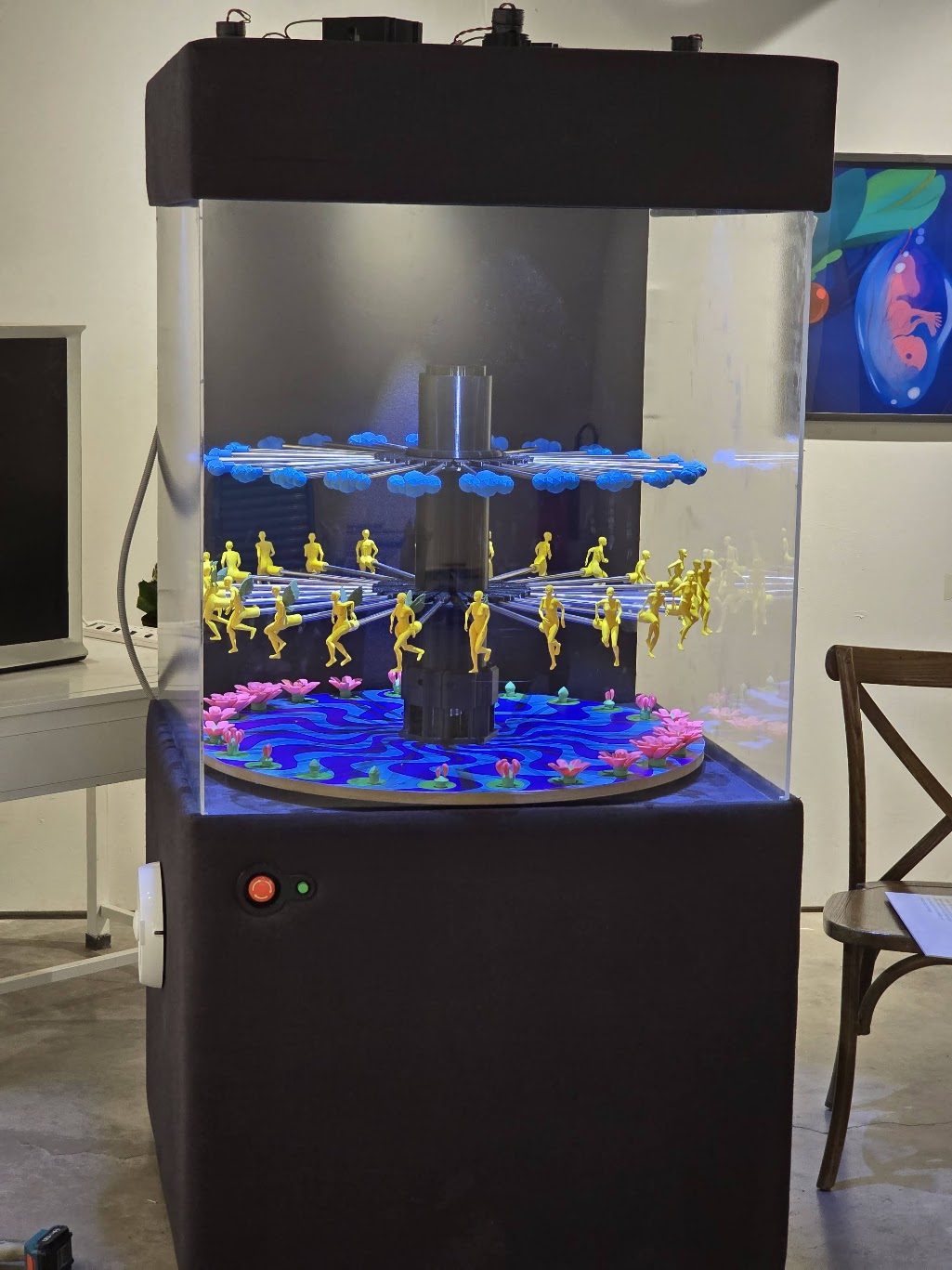 The finished zoetrope standing in the gallery — a dark upholstered plinth with a clear acrylic vitrine above it, the three lit layers turning inside, power and E-stop buttons on the front of the plinth