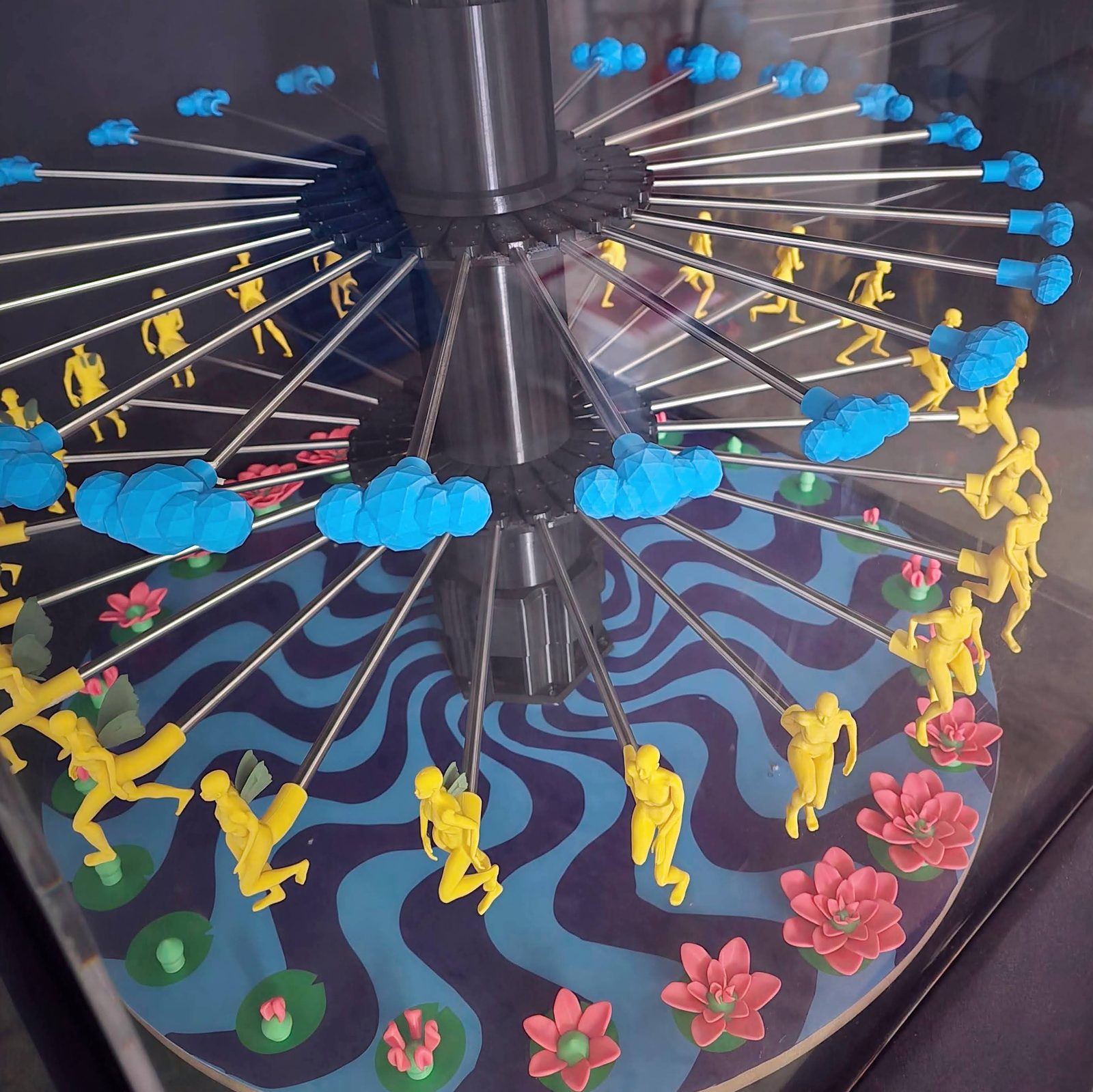 Close view down into the assembled zoetrope — blue clouds on the top ring, yellow human forms on the middle ring, pink lotuses on the painted platter below, all on radial steel spokes off the black centre tower
