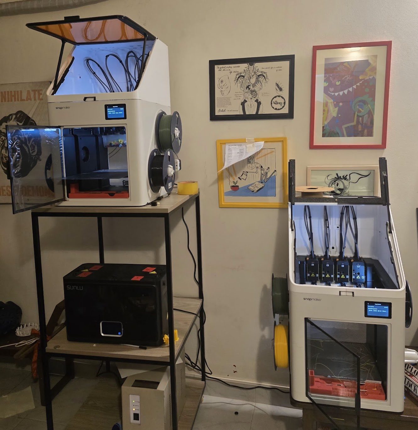 Two Snapmaker U1 3D printers, one on a steel shelf unit and one standing beside it with its four-tool station open, next to a Sunlu S4 Pro filament dryer and spools of filament