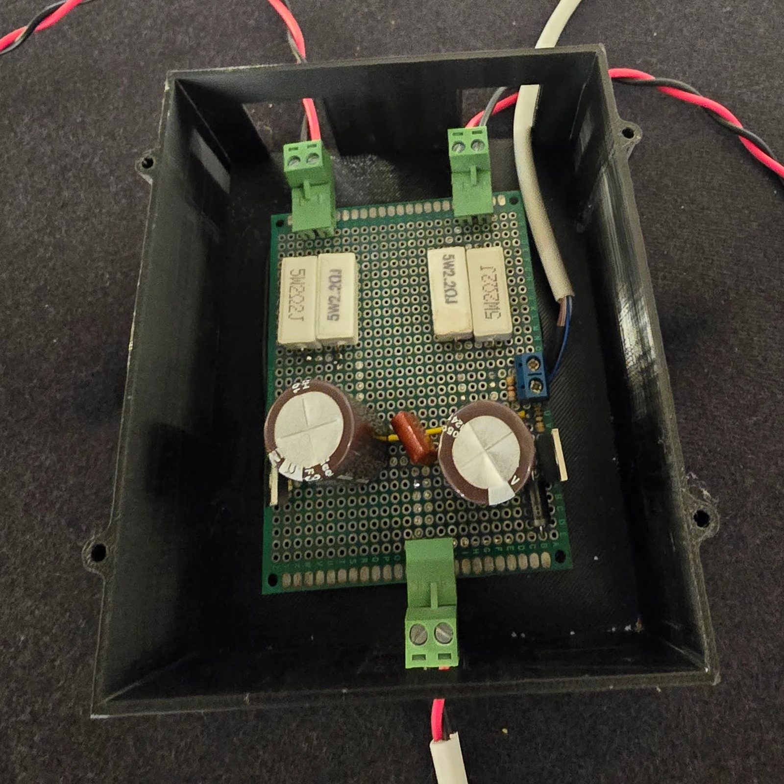 The driver board built on perfboard inside its printed enclosure with the lid off — four white ceramic ballast resistors in two pairs, two large electrolytic bulk capacitors, the bleed resistor between them, three green screw terminals and one blue, and twisted red-black pairs leaving the box
